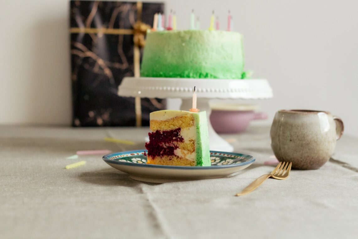 Stück einer grünen Torte mit roter Himbeerfüllung und Kerze auf einem Teller, dahinter die ganze Torte mit Geburtstagskerzen